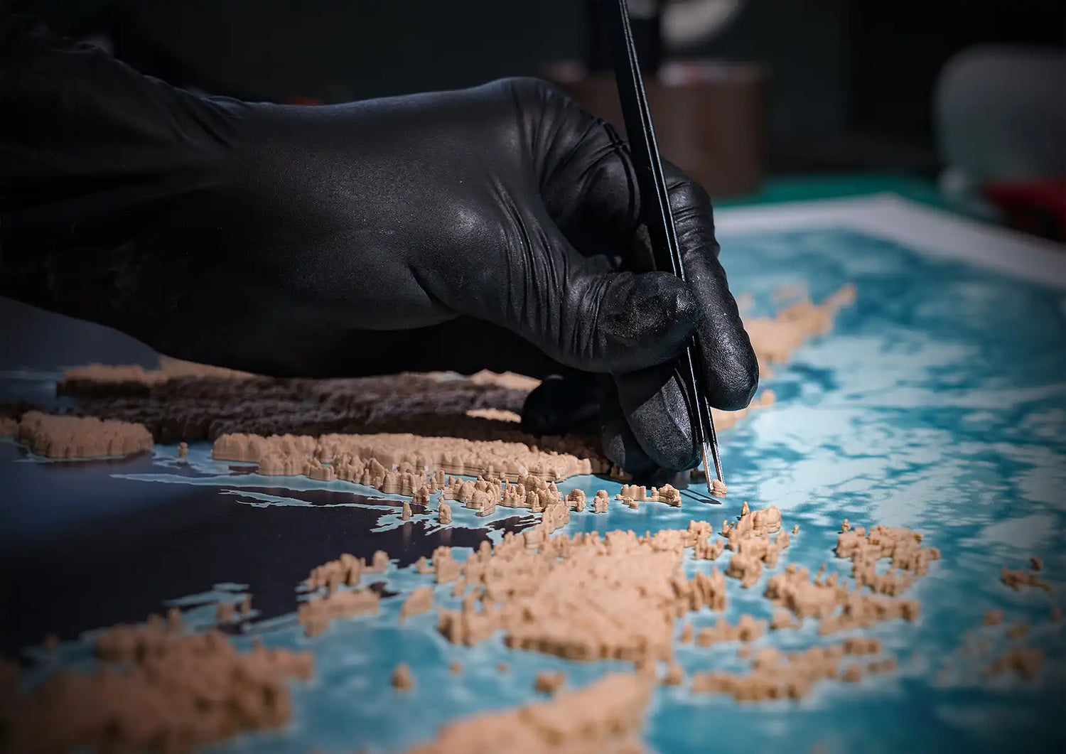 Person wearing black gloves carefully placing small parts on a 3 topographic map of many islands - TerraFrames