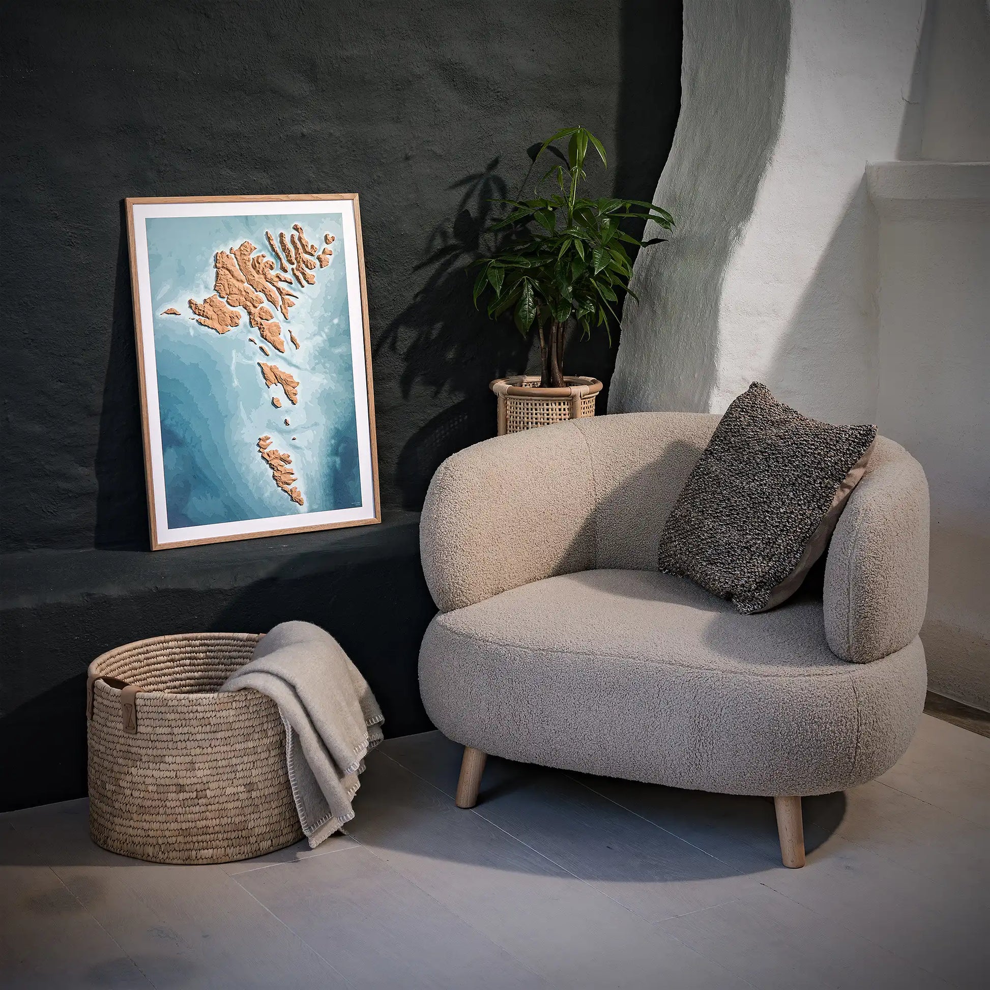 Framed 3D topographic map of the Faroe Islands, in a minimalist room with a beige armchair, a woven basket, a plant and a black wall – TerraFrames