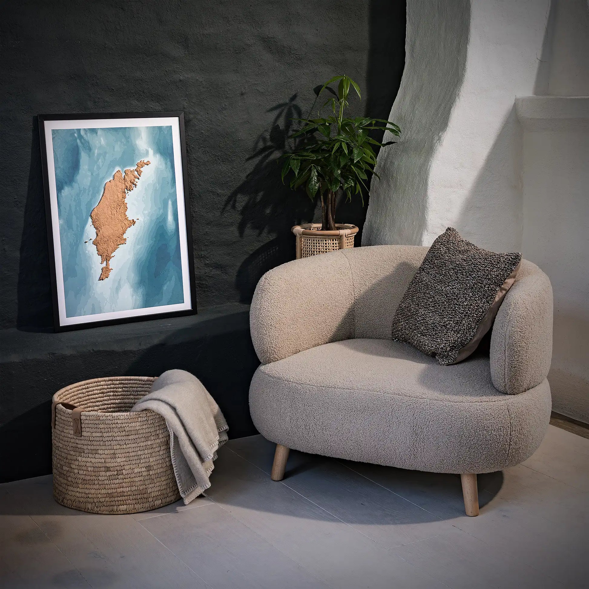 Framed 3D topographic map of Gotland, in a minimalist room with a beige armchair, a woven basket, a plant and a black wall – TerraFrames