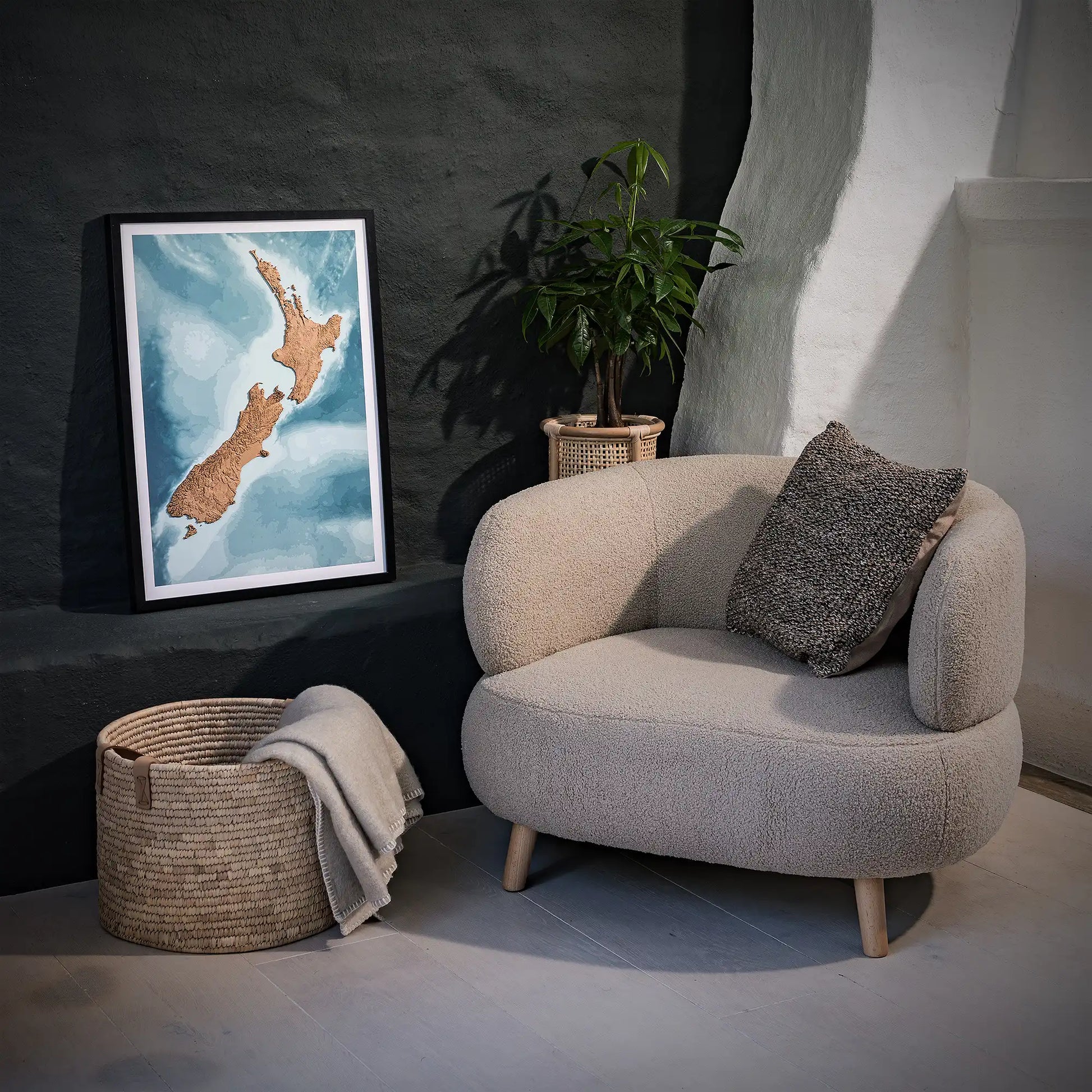 Framed 3D topographic map of New Zealand, in a minimalist room with a beige armchair, a woven basket, a plant and a black wall – TerraFrames