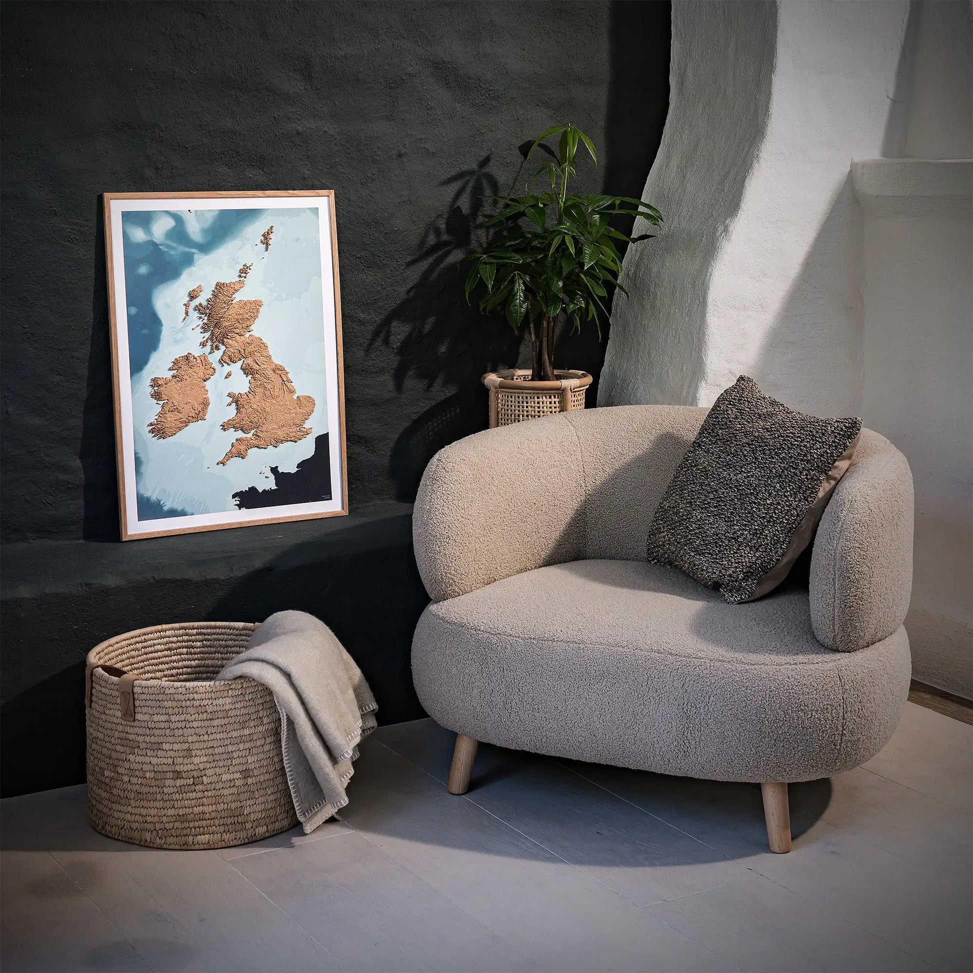 Framed 3D topographic map of Scotland, England and Ireland, in a minimalist room with a beige armchair, a woven basket, a plant and a black wall – TerraFrames